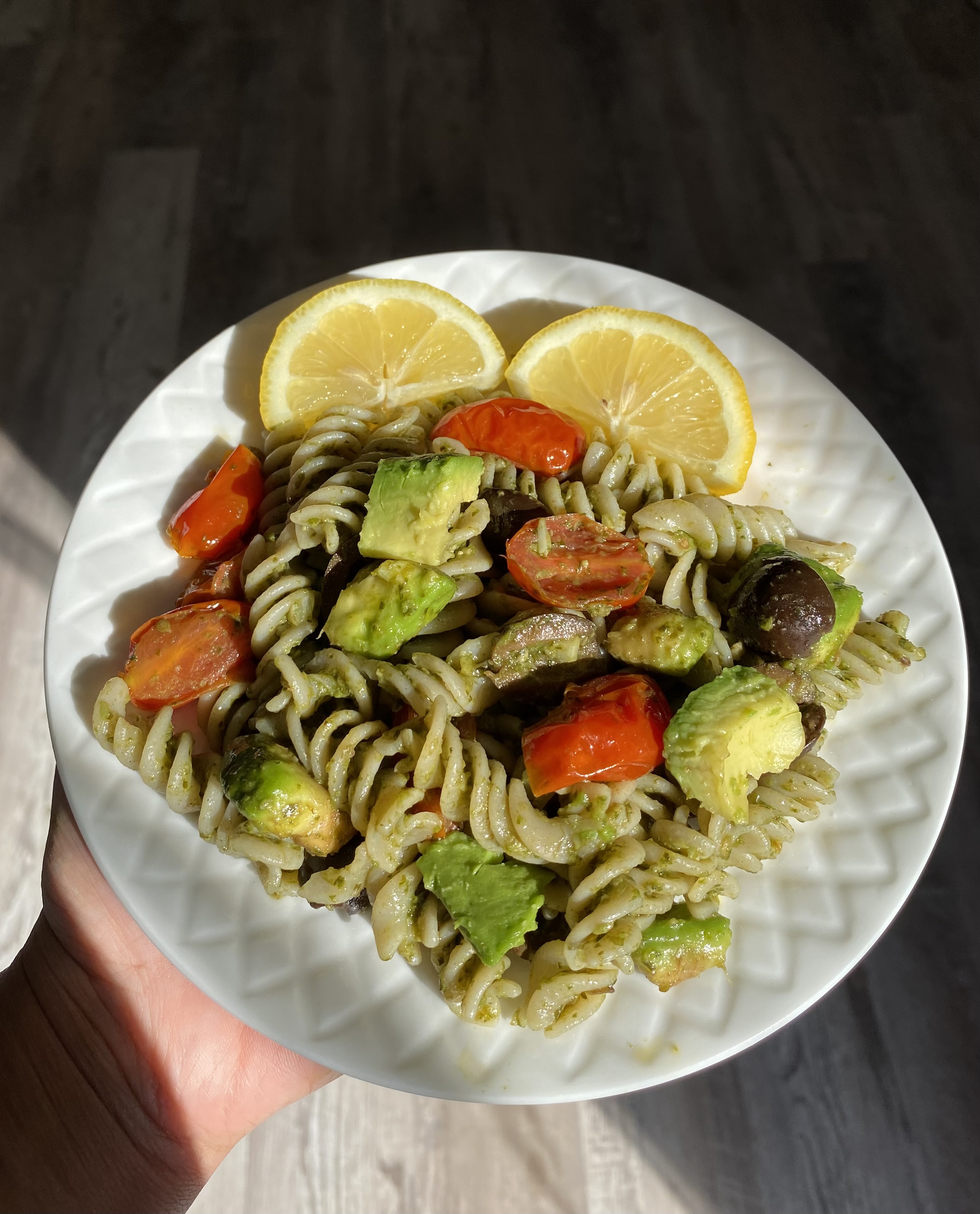 Gluten Free Vegan Avocado Pesto Pasta with sautéed onions, tomatoes, and black olives. Served on a white plate with 2 half lemon slices.